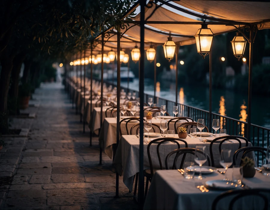 Lantern-lit riverside restaurant terrace with linen-set tables along the San Antonio River at dusk